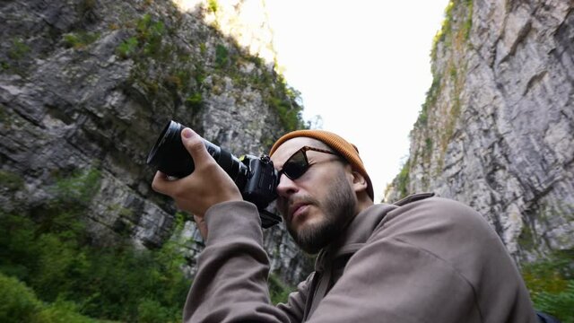 Young bearded tourist in sun glasses with camera taking pictures of wild mountain rocky nature. Adult traveler photographer on vacation trip in highland forest among rocks. High land natural tourism.