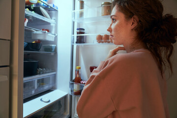 Undecided young caucasian woman checking fridge for some food at night - obrazy, fototapety, plakaty