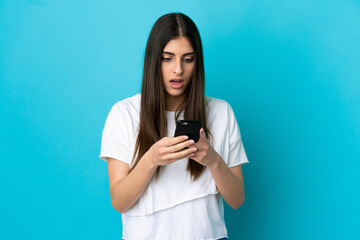 Young caucasian woman isolated on blue background looking at the camera while using the mobile with surprised expression
