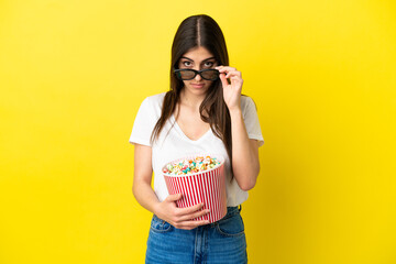 Young caucasian woman isolated on yellow background surprised with 3d glasses and holding a big bucket of popcorns
