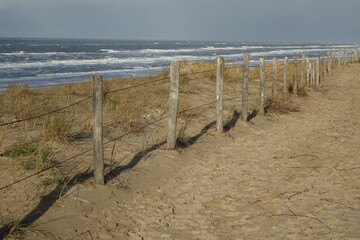 Sandiger Dünenpfad mit Zaun am Nordseestrand mit rollenden Wellen an einem sonnigen, stürmischen Wintermorgen, Egmond aan Zee, Nordholland, Niederlande