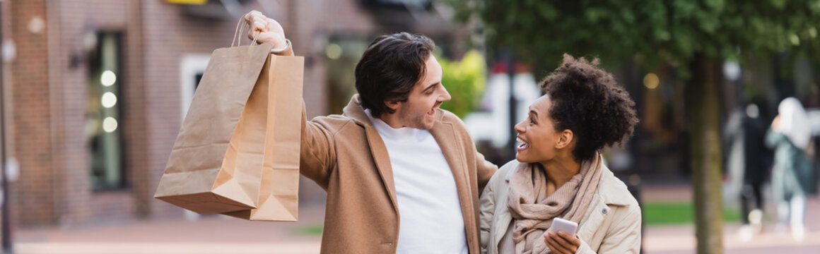 amazed african american woman with smartphone looking at happy boyfriend holding shopping bags, banner.