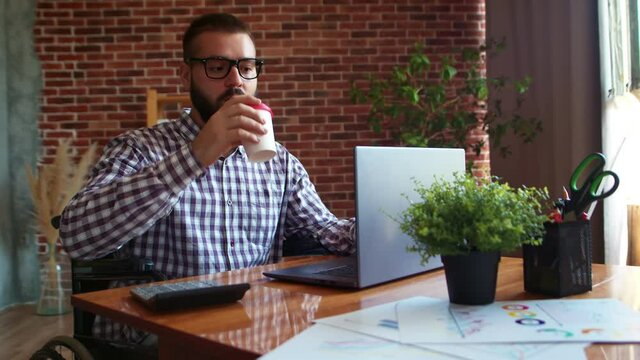 Man wheelchair user is working on laptop, drinking coffee, looking ahead and smiling. Hipster businessman took break from work and drinks coffee. Concept of possible work for people with disabilities
