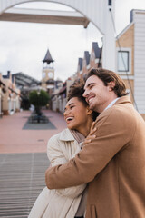 Fototapeta premium joyful multiethnic couple in coats hugging near shopping center.
