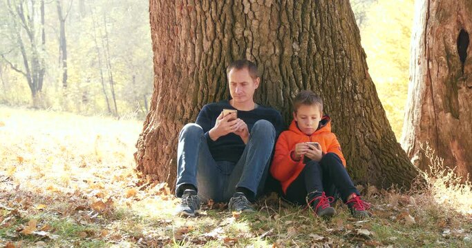 Father And Son Rests Under the Huge Oak in National Park While Hiking in Autumn. Family Use Smartphones in Nature and Talk with Each Other