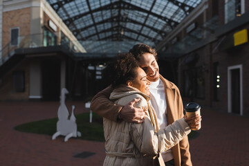 cheerful man hugging happy african american woman with paper cup in shopping mall.