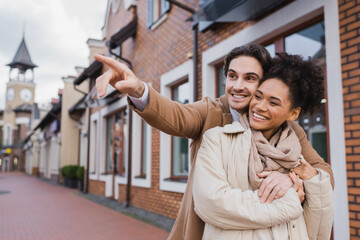 Fototapeta premium happy man pointing away with finger while hugging african american girlfriend outdoors.