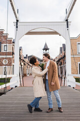 Fototapeta premium full length of smiling multiethnic couple in coats hugging near shopping center.