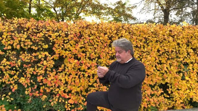 Man standing outside texts on the phone. In the background the colorful foliage shows that it is during autumn season.