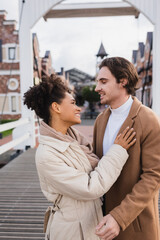 smiling multiethnic couple in coats embracing near shopping center.