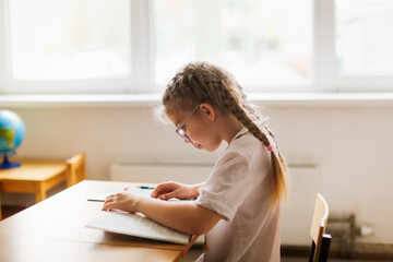 cute girl child with glasses reads a book at a desk at school, learning concept, private small...