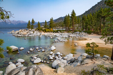 Very rocky shoreline of Lake Tahoe with clear, still blue water, mountains and trees against a blue...