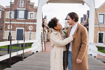 smiling multiethnic couple in coats hugging near shopping center.