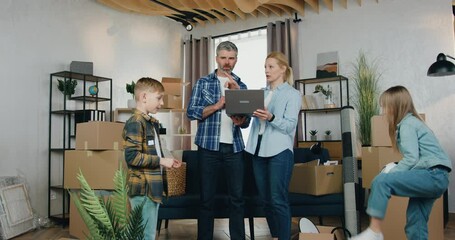 Adorable skilful satisfied adult couple discussing ideas of room design using computer program while their cheerful playful 10-aged son and daughter having fun with ball