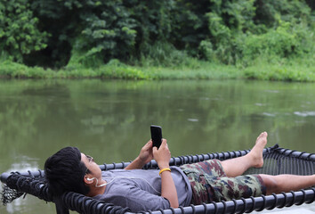 black haired man, ethnicity unknown lying on the outdoor net using a mobile phone by the river to listen to music Take a vacation with relaxation and peace of mind. Ideas for holiday activities