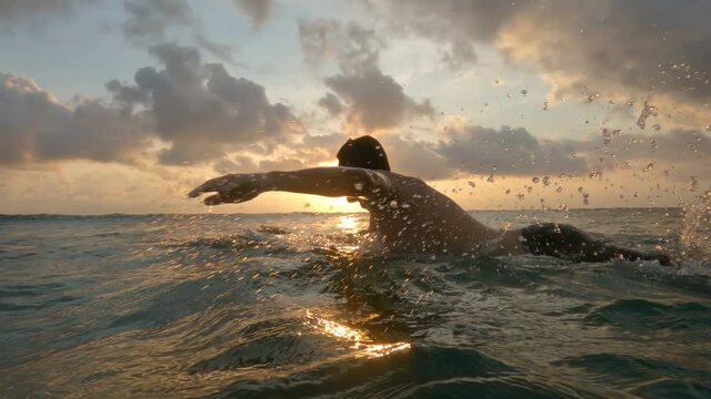A handsome young surfer in shorts swims on a white surfboard through the waves against the backdrop of a wonderful sunset with a bright sun and thick clouds. A man is surfing in the ocean. Slow motion