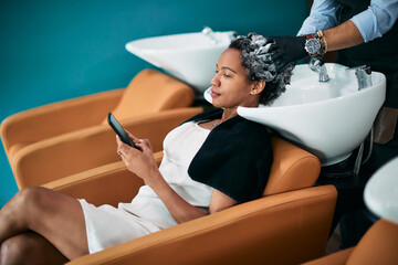 Black woman reads text message on mobile phone while washing hair at hairdresser's.