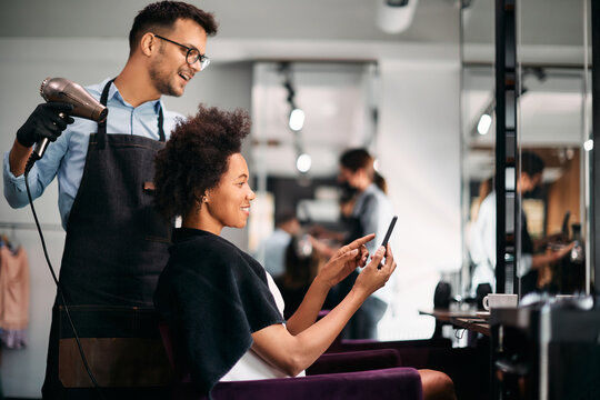 Happy African American woman shows something on smart phone to her hairdresser while getting her hair styled at salon.