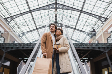 low angle view of happy multiethnic couple with paper cups and shopping bags on escalator in mall.