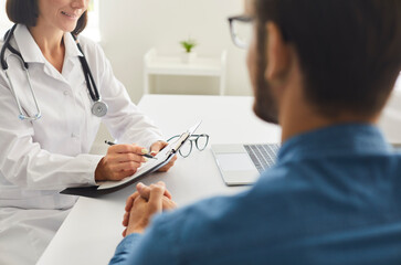 Woman who works as physician sitting at desk, talking to young man, writing down patient's...