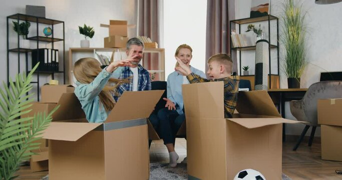 Likable smiling happy adult couple talking on the couch between themselves when their two kids jumping out carton boxes with joy,family in new apartment after relocation