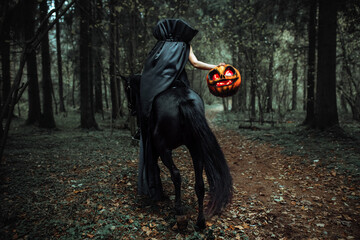 the rider is sitting astride a horse. a girl in a long black raincoat with a pumpkin mask with...