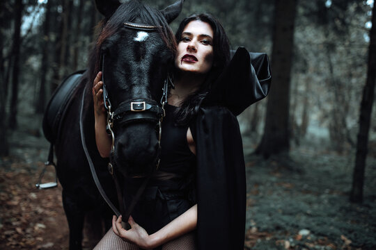 mask in the form of a pumpkin with burning eyes. creepy girl costume for halloween. a rider in a long black cloak holds a horse by the bridle. cosplay of a gloomy image in the autumn forest