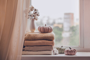 A stack of warm knitted sweaters on the windowsill with pumpkins and a vase of cotton plant in pastel shades