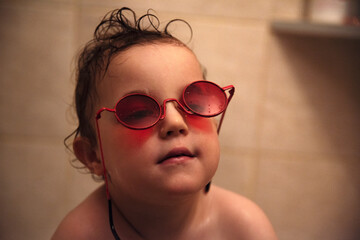 Mały chłopiec w popsutych czerwonych okularach przeciwsłonecznych, siedzący w wannie podczas...