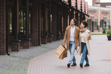 positive multiethnic couple in coats holding hands while walking with purchases near mall.