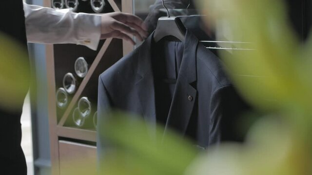 An elegant man removes a black jacket from a hanger hanging by the window in the apartment.