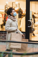 young african american woman in medical mask walking with purchases in shopping center.