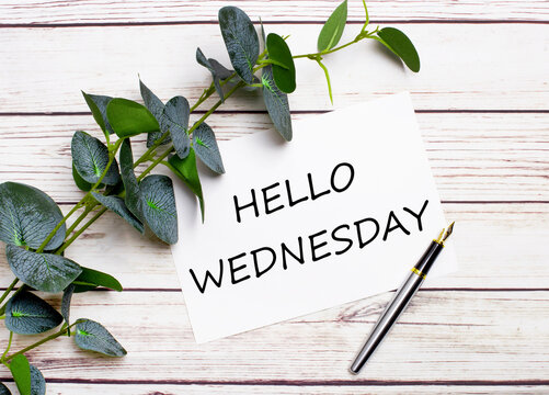 On a light wooden table, there is a eucalyptus branch, a fountain pen and a sheet of paper with the text HELLO WEDNESDAY