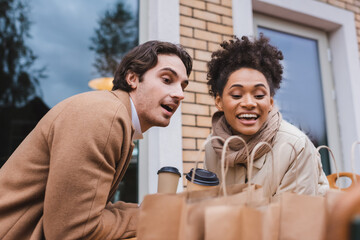 Obraz premium joyful interracial couple in coats holding paper cups and looking at blurred shopping bags.