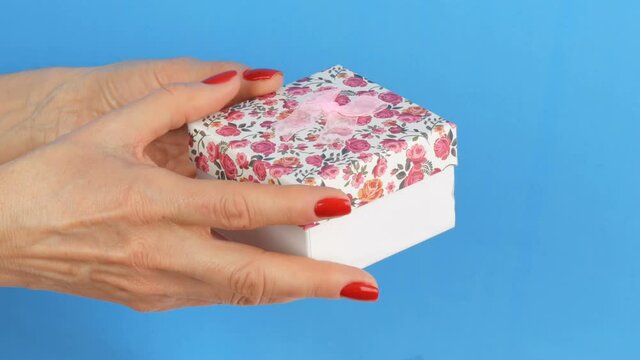 A small gift box with a floral print and a pink bow that is held by the hands of woman whose nails are painted with red nail polish on a blue background