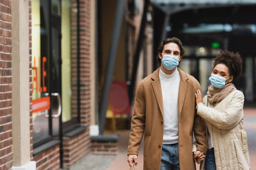 young multiethnic couple in medical masks holding hands while walking in shopping mall.