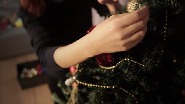 Close up on her hand. A girl prepares a New Year tree. She chooses a Christmas toy in a large black box and hangs a large golden ribbed and red ball on a Xmas tree is decorated with red and gold balls