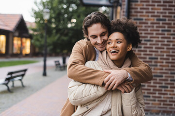 happy man embracing amazed african american girlfriend near building outdoors.