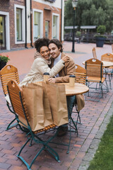 happy interracial couple embracing near paper cups and shopping bags in outdoor cafe.