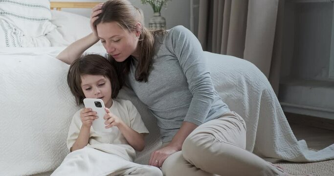Confident mother learning cute little son use smartphone surfing internet helpful application and call. Modern female and kid boy studying mobile phone digital technology device telephone addiction