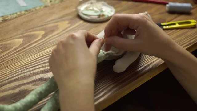 Women's hands sews details of rag doll on wooden table. Handmade toy made of linen, sintepon and synthetics. Process of sewing legs to body of doll. Craft diy. Close-up.