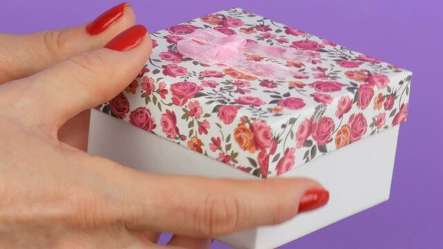 Small gift box with a floral print and a pink bow that is held by the hands of woman whose nails are painted with red nail polish on a purple background