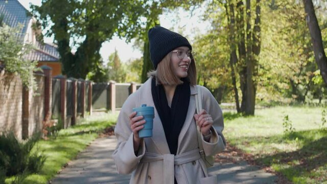 Young girl walks in the green neighborhood on a sunny autumn day, smiling and holding an eco cup. Beautiful girl walking in the park. Front view. High quality 4k footage