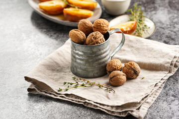 A metal mug with whole brown walnuts on a beige colored linen cloth, caramelized pears and thyme branches on a grey concrete surface