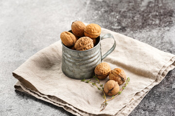 A metal mug with whole brown walnuts on a beige colored linen cloth on a grey concrete surface