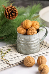 A metal mug with whole brown walnuts on a checkered linen cloth and a christmas fair-tree branch on a white table surface