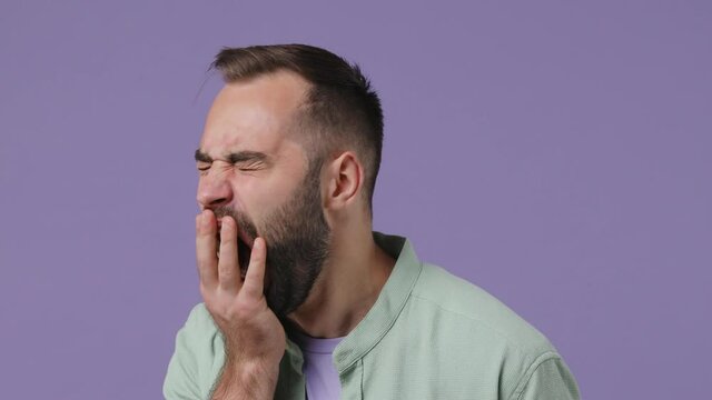 Close up tired sad young bearded man 20s wear mint shirt did not get enough sleep last night after party and barely got up in the morning yawn isolated on plain light purple background studio portrait