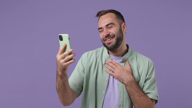 Fun young bearded man 20s wears mint shirt get video call using mobile cell phone do selfie talk conduct pleasant conversation greet with hand isolated on plain light purple background studio portrait