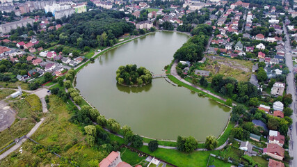 Fototapeta na wymiar View of the pond in the park in Kolomyia. Island on a city lake. Roofs of residential buildings. Ukraine, Europe
