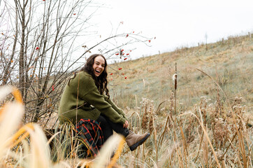 Young laughing brunette woman is sitting on a stump in autumn forest among dry grass, looking at the camera.